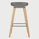 Vista frontal del banco alto Perique en color gris, destacando su silueta minimalista y el contraste entre el asiento sintético y las patas de madera clara.