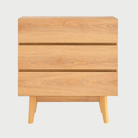 Vista frontal de la cómoda Meztli B en acabado madera beige clara, que cuenta con tres cajones de frentes lisos y apertura mediante bisel superior, apoyada sobre patas de madera inclinadas.