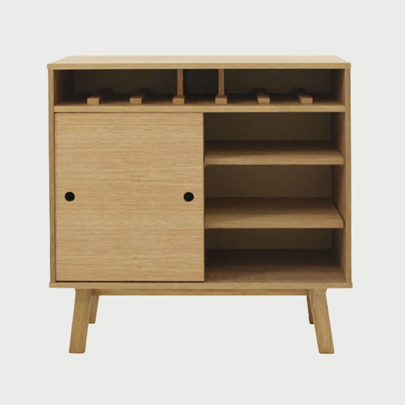 Vista frontal del mueble de bar Avilés en acabado beige encino, con una sección superior para copas, una puerta deslizable con tiradores circulares y estantería abierta lateral sobre patas inclinadas.