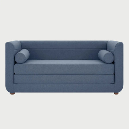 Vista frontal del sofá cama Basil en tela azul, con diseño de respaldo bajo, dos cojines cilíndricos laterales y patas de madera en acabado nogal oscuro.