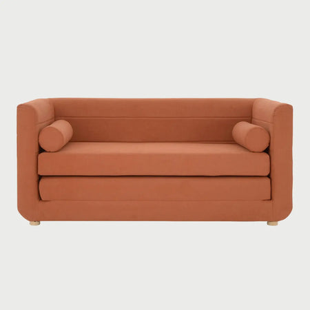 Vista frontal del sofá cama Basil en color café claro (beige), destacando los cojines cilíndricos laterales y las patas de madera natural en acabado barnizado.