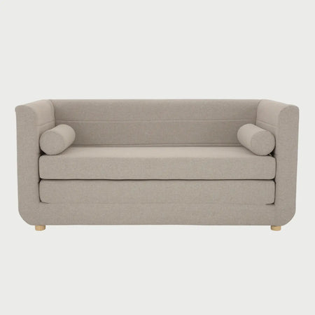 Vista frontal del sofá cama Basil de GAIA en tela amarilla, con un diseño de respaldo bajo y brazos integrados de altura uniforme, asiento de doble capa y dos cojines decorativos cilíndricos en los extremos.