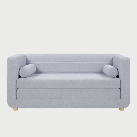 Vista frontal del sofá cama Basil en color azul, destacando su silueta moderna y acogedora con cojines de tubo laterales y patas de madera clara de perfil bajo.