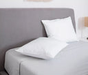 Escena de dormitorio moderno que muestra un set de almohadas Brilliance Premium en color blanco sobre una cama con sábanas grises. La composición resalta los diferentes niveles de firmeza y el acabado de lujo de la colección, complementando un ambiente de descanso sereno y minimalista.