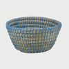 Vista frontal de la canasta chica Damu; presenta una forma circular de base amplia tejida con fibras naturales de paja, reforzada con un tramado de hilo color azul vibrante que crea un patrón LINEAs verticales envolventes.