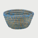 Vista frontal de la canasta chica Damu; presenta una forma circular de base amplia tejida con fibras naturales de paja, reforzada con un tramado de hilo color azul vibrante que crea un patrón LINEAs verticales envolventes.