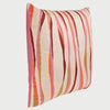 Vista lateral en ángulo del cojín cuadrado Serpentina de 50 x 50 cm; se observa su perfil con un relleno generoso que le otorga un volumen suave y redondeado. El costado muestra un acabado en rosa sólido, mientras que la cara principal presenta un patrón LINEAs verticales irregulares y onduladas en tonos rosa, naranja, borgoña y ocre sobre un fondo crema.