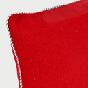 Acercamiento al cojín Víspera en color rojo; se aprecia la textura densa y cálida de la lana natural junto con el detalle del ribete perimetral trenzado en hilos blanco y negro, que aporta un contraste gráfico y refuerza la estructura del borde.
