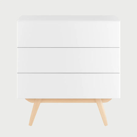 Vista frontal de la cómoda Cuauhtémoc en color blanco brillante, con tres cajones de frentes biselados sin tiradores externos y una base de madera clara con patas angulares.