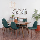 Un comedor de estilo moderno con la mesa Talitha de 200 cm en color nogal como pieza central, rodeada por seis sillas tapizadas en color verde oscuro; la escena incluye un bufetero al fondo con espejos decorativos, lámparas colgantes geométricas y una planta de interior grande, todo sobre un piso de madera laminada.