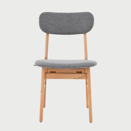 Vista frontal de la silla Marcela con estructura de madera en acabado encino natural y asiento acolchado tapizado en tela gris; destaca su respaldo ovalado horizontal y patas cilíndricas.
