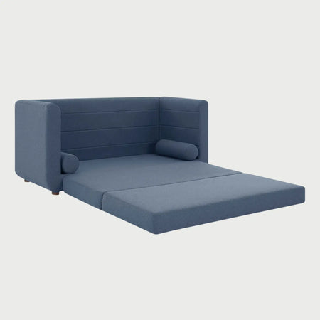 Vista lateral del sofá cama Basil azul, resaltando el perfil redondeado de sus brazos y la elegancia de su base elevada sobre patas de madera acabado nogal.