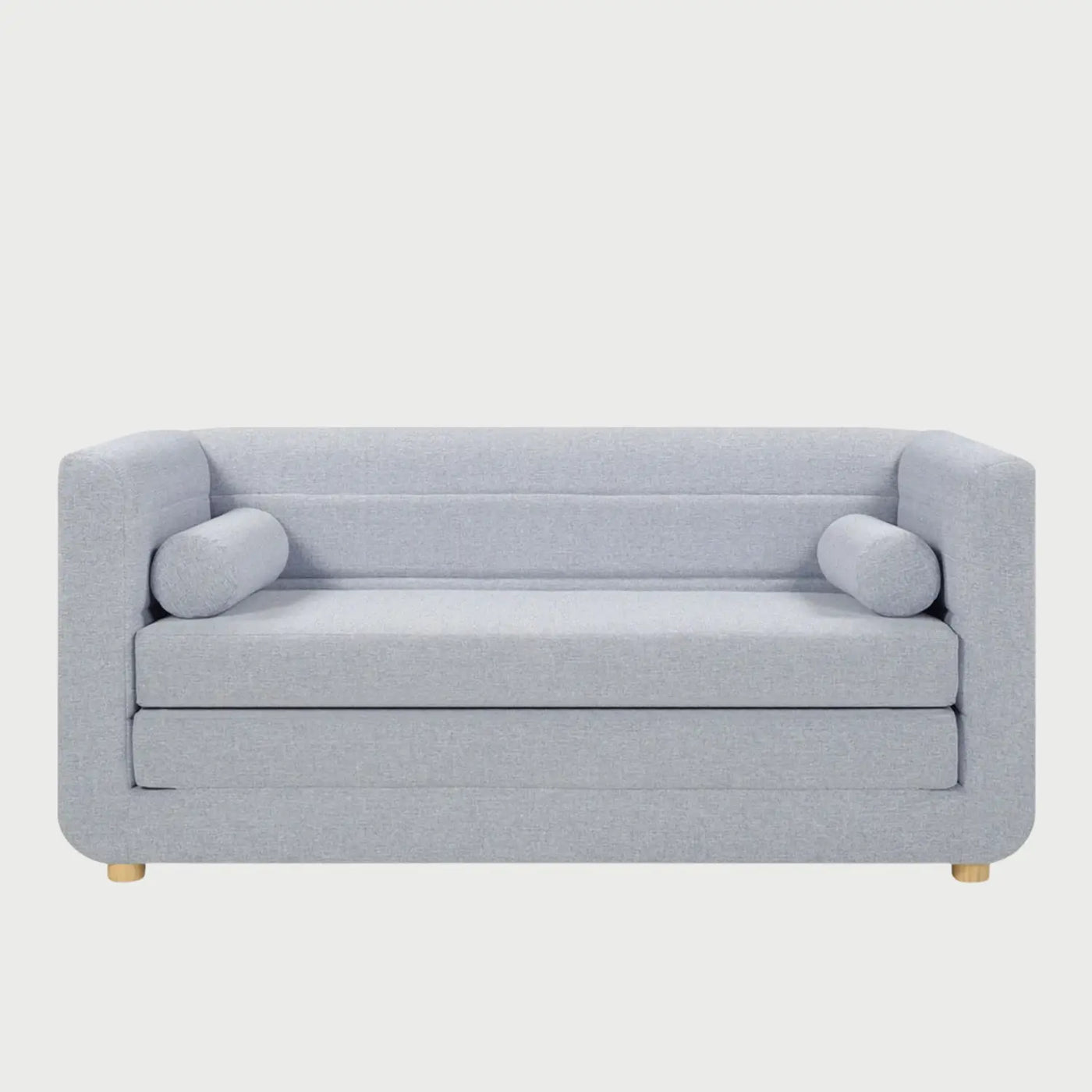 Vista frontal del sofá cama Basil en color azul, destacando su silueta moderna y acogedora con cojines de tubo laterales y patas de madera clara de perfil bajo.