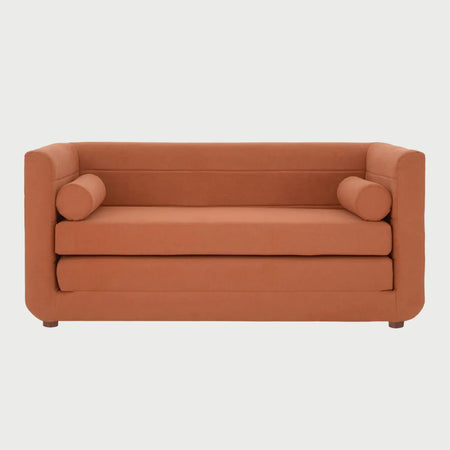 Vista frontal del sofá cama Basil en color naranja vibrante, resaltando sus cojines de tubo laterales y el contraste cálido con las patas de madera oscura.