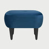 Vista frontal del taburete Tavari tapizado en terciopelo azul, con un asiento de silueta ovalada, un detalle de botón central tipo capitoné y cuatro patas cónicas de madera en acabado negro.