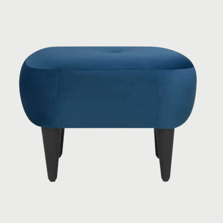 Vista frontal del taburete Tavari tapizado en terciopelo azul, con un asiento de silueta ovalada, un detalle de botón central tipo capitoné y cuatro patas cónicas de madera en acabado negro.